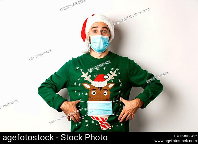 Concept of covid-19 and Christmas holidays. Funny man put on face mask on his sweater deer, standing over white background
