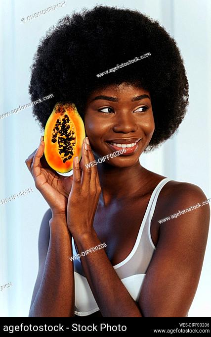 Smiling female model in white bra looking away while holding slice of papaya against wall