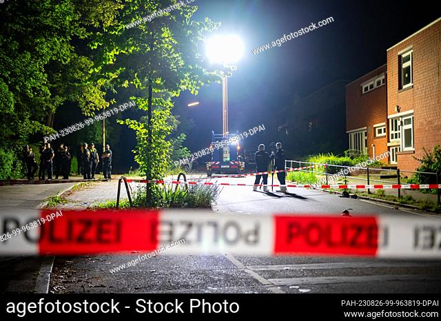 26 August 2023, Hamburg: A light pole illuminates the cordoned-off crime scene while forensic technicians work with a 3D scanner and other police officers...