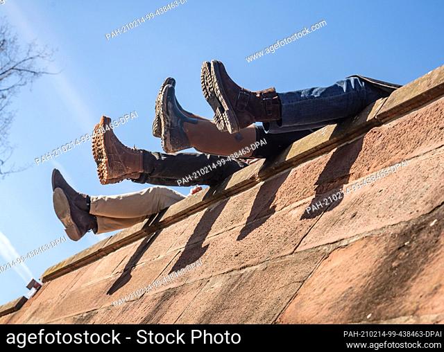 14 February 2021, Hessen, Frankfurt/Main: Four people dangle their legs from the high wall on the Mainkai in the afternoon as they sit together in the sunshine