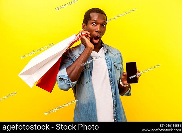 Wow, amazing mobile app for online shopping. Portrait of happy surprised man holding packages and showing smartphone with amazed shocked expression
