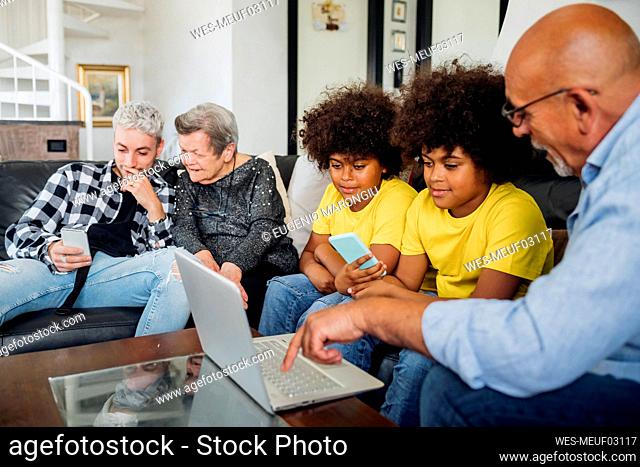 Twin boys with grandfather using laptop while young man using smart phone sitting by great grandmother in living room