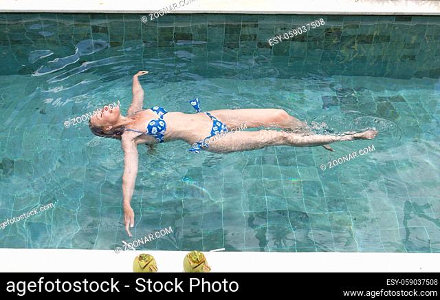 The happy slender young woman lies on a water surface in the pool and has a rest