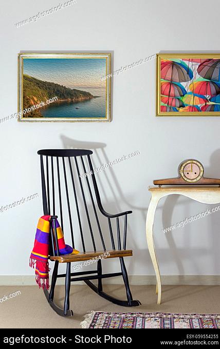 Interior shot of vintage rocking chair and antique desktop clock on old style vintage table on background of off white wall with two hanged paintings including...