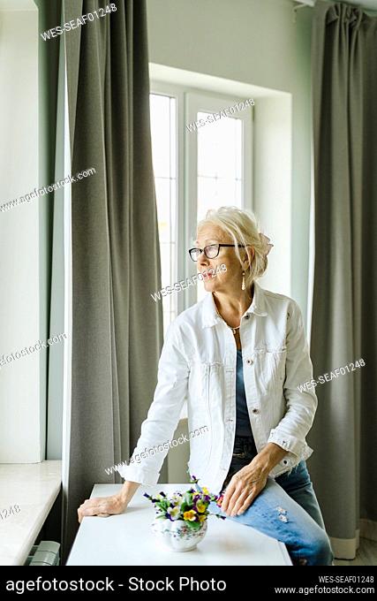 Thoughtful woman sitting on table near curtain at home