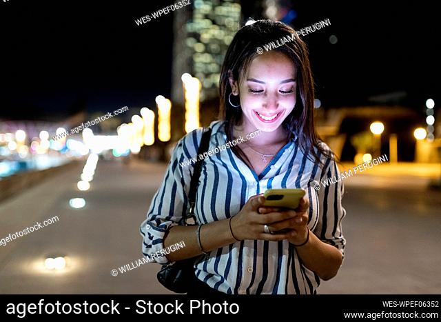 Smiling young woman text messaging through mobile phone at night