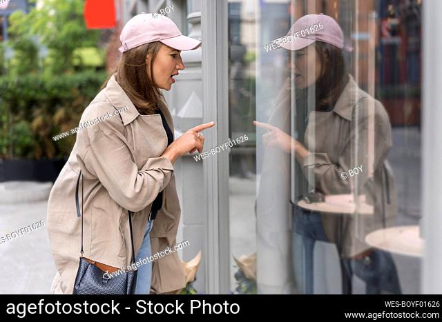 Woman wearing cap pointing at reflection on glass door in city