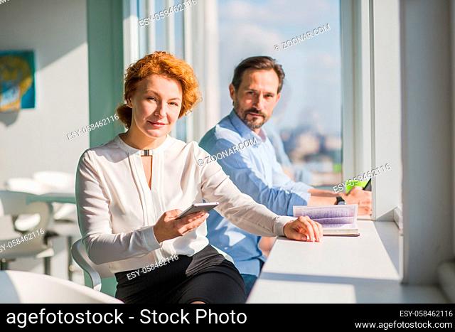 Businesswoman using mobile or smart phone in office interior while looking at camera. Her colleague businessman looking at her and having rest or break