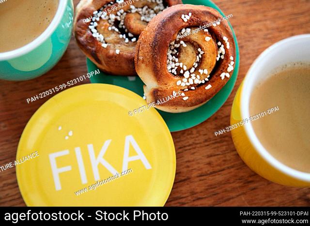 ILLUSTRATION - 14 March 2022, Denmark, Kopenhagen: A saucer with the imprint ""Fika"" lies next to two cinnamon buns and two mugs with coffee
