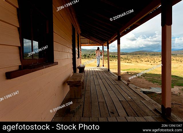 Woman standing on verandah of rustic timber homestead with scenic views in the snowy high plains. A storm is brewing out over the ranges