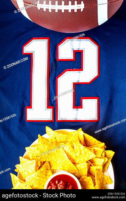 An American football with organic nacho chips and mild salsa on a white blue football jersey with the 12 number on vertical view