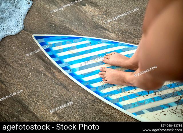 Macro photo of the female legs which are standing on the light surfboard with gray-cyan stripes on the beach. Closeup. Horizontal