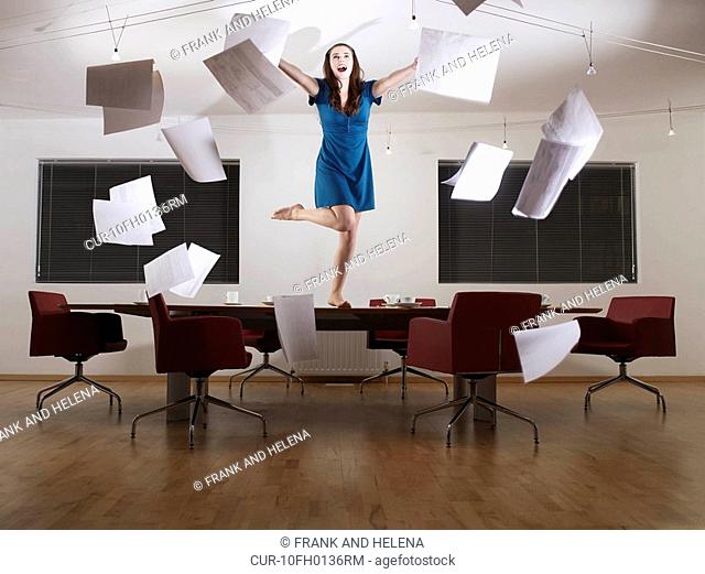 Young woman throwing papers in the air, while standing on conference table, dancing and posing. She is smiling