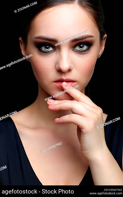 Beauty portrait of young woman with hand near face on black background. Brunette girl with evening female makeup and black dess touches lips with fingers