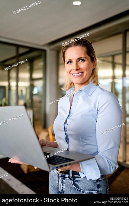 Smiling businesswoman with laptop standing at workplace