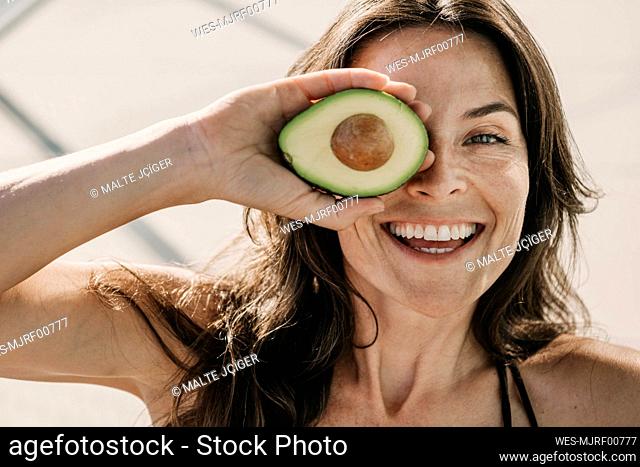Happy mature woman covering eye with halved avocado