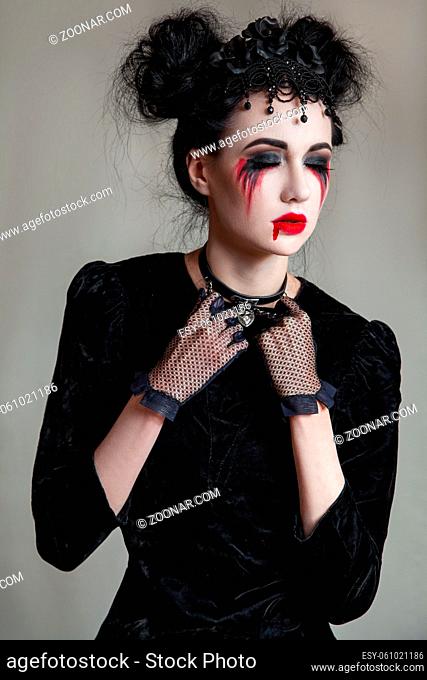 Young beautiful gothic woman with white skin and red lips with bloody drops wearing black collar with spikes. Red smokey eyes. Halloween makeup