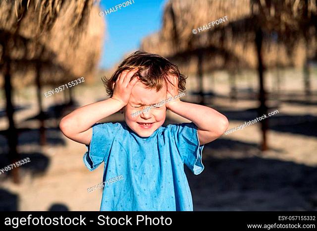 A small girl with headache walking among straw parasols outdoors on beach, eyes closed