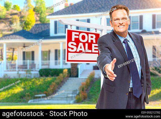 Smiling Male Agent Reaching for a Hand Shake in Front of Beautiful House and For Sale Real Estate Sign