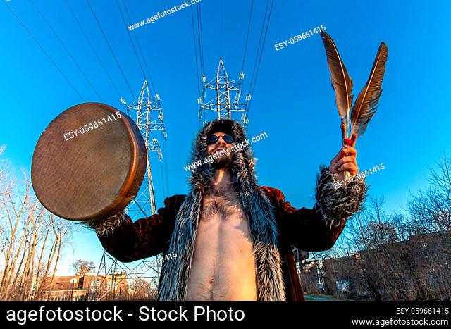 A low angle and front view of a happy medicine man holding sacred drum and symbolic feathers as he seeks inspiration in city, shirtless beneath clear blue sky