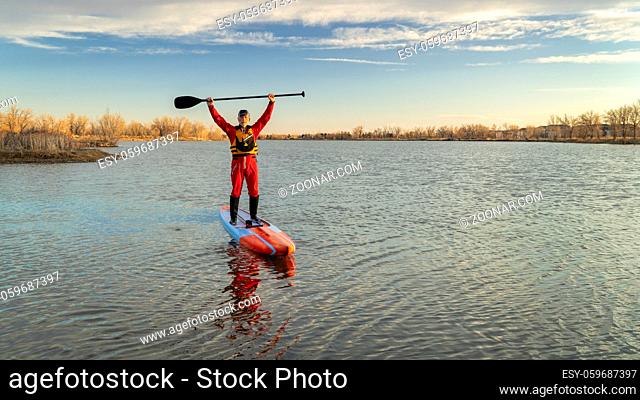 senior male paddler in a drysuit and life jacket is paddling a long racing stand up paddleboard on a lake in Colorado, winter or early spring scenery