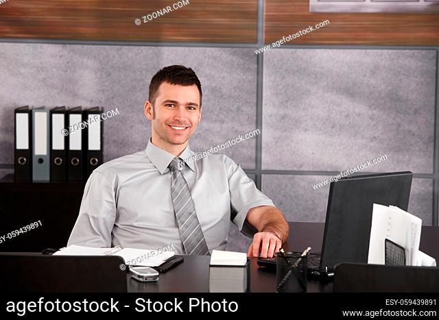 Portrait of casual businessman sitting at desk wearing short sleeved shirt, smiling. Copyspace on right
