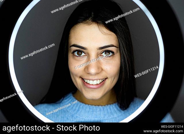 Young woman with illuminated circular camera flash smiling while standing against gray background