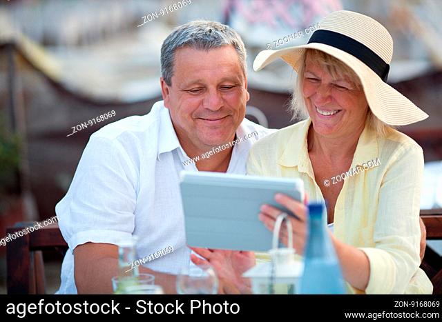 Smiling happy relaxed middle-aged couple using a tablet together on vacation as they sit at a table in a cafeteria enjoying refreshments
