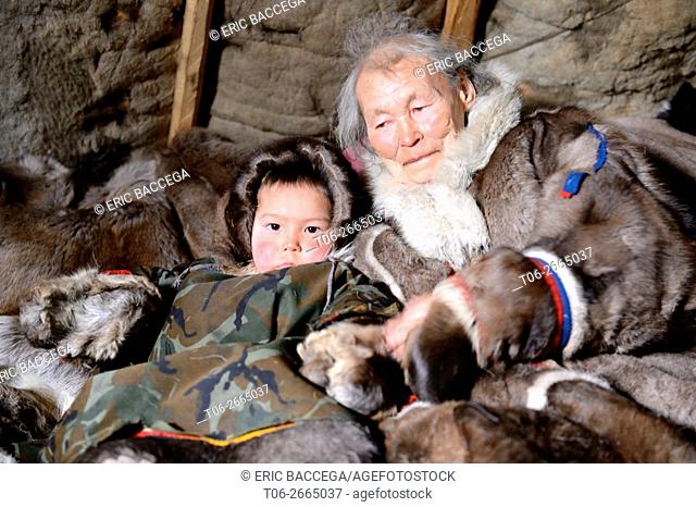 A Nenets older woman with her grandson. Both are dressed in their winter coats, made with reindeer skin and sitting in their tent / Chum, Yar-Sale district