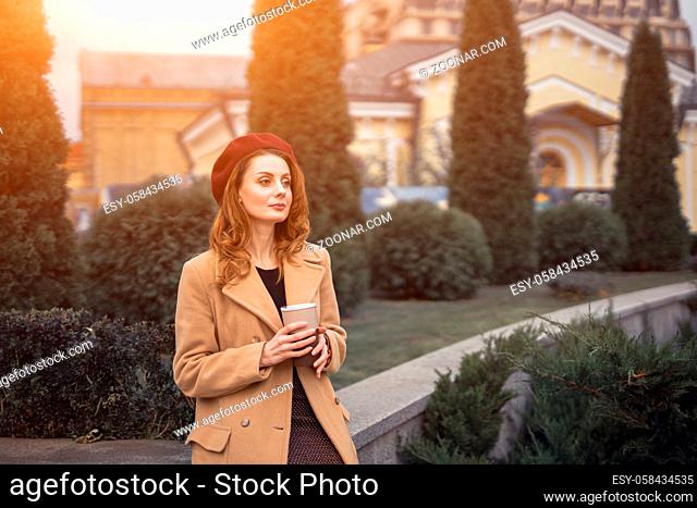 Pensive beautiful young woman holding a cup of coffee on the street female fashion. Portrait of stylish young woman wearing autumn coat and red beret outdoors