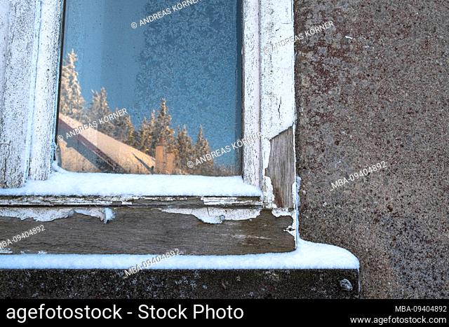 Snow-covered, old windowpane with frosted flowers and reflection of a sunny, wintry landscape and hut in gray facade in Vosges, France