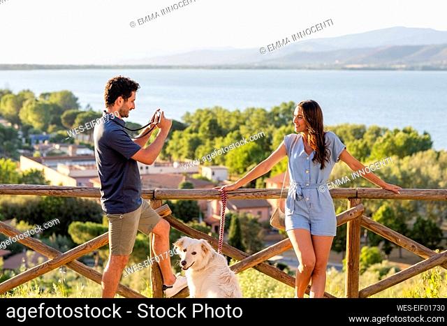 Man photographing woman through camera while standing with dog by railing