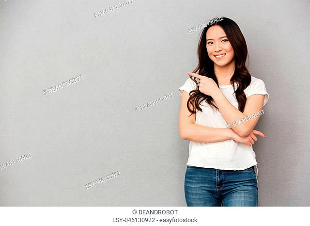 Photo of asian affable woman in casual t-shirt and jeans posing on camera and pointing finger aside on copyspace isolated over gray background