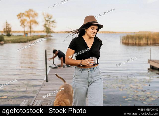 Smiling woman holding wineglass while walking with dog and man in background on pier