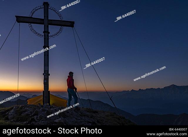 Sunrise over Ammergau mountains, in the foreground summit cross of the Geierkopf with mountaineer and tent, Reutte, Ammergau Alps, Tyrol, Austria, Europe