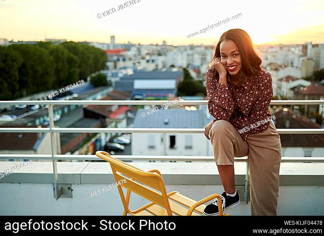 Smiling young woman with hand on chin standing by chair in balcony