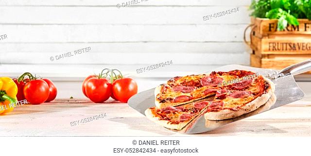Panorama banner with a traditional Italian salami pizza served on a metal paddle with a slice removed and fresh ingredients and herbs on a rustic white wood...