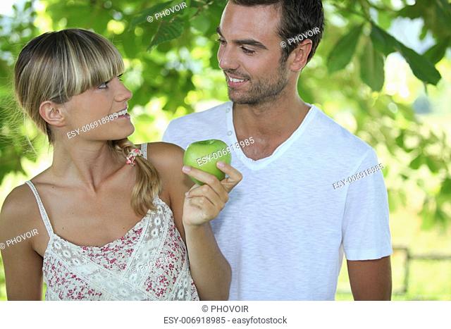 a blonde woman and a man are looking at each other, the woman is taking an apple, the background is forestry