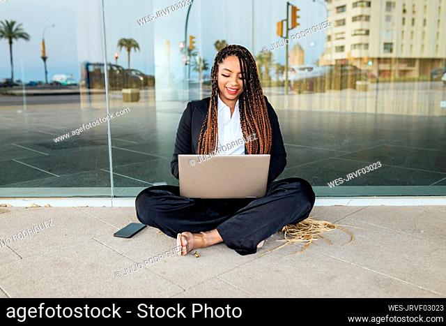 Happy businesswoman sitting and working on laptop outside glass wall