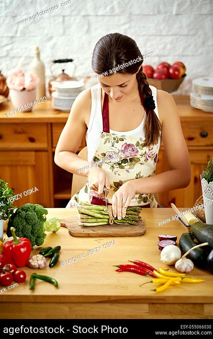 beautiful young woman, brunette slicing asparagus in the kitchen at a table full of organic vegetables