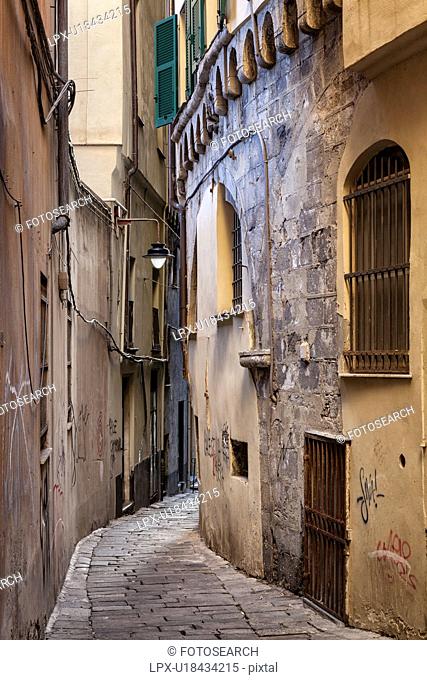 Genoa Centro Storico: narrow cobbled alleyway with medieval stone facade visible under peeling paint and graffitti covered stucco, Genova, Italy