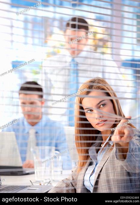 Attractive young businesswoman looking through blind in bright office