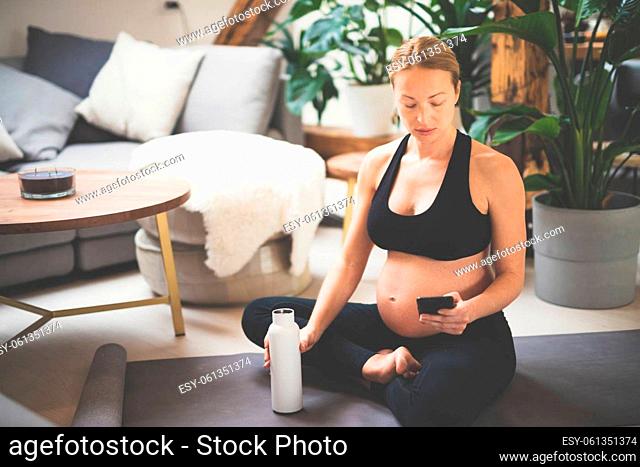 Young happy and cheerful beautiful pregnant woman taking a break, hydrating, drinking water from the botle after home well being workout program