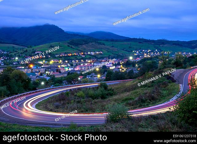 Cloudy evening on a mountain road in the Ukrainian Carpathians. Traces of headlights of fast moving cars. Small village shining with lights in the valley