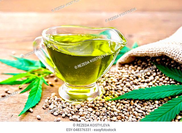 Hemp oil in a glass sauce-pot with grain in a bag, leaves and stalks of cannabis on a wooden board background