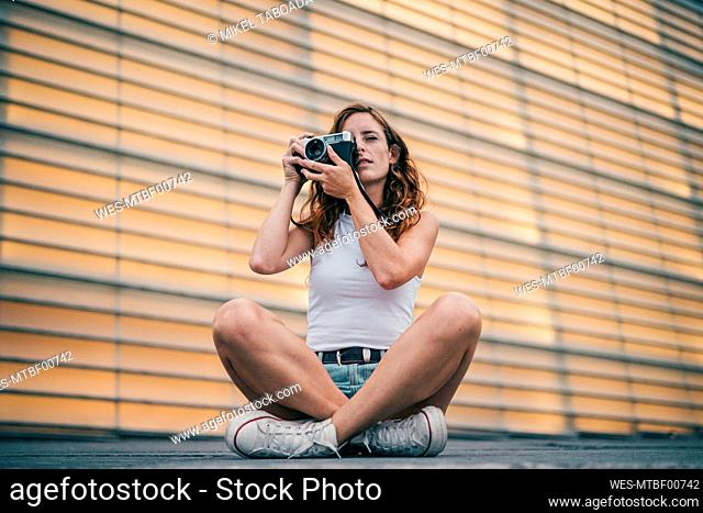Young woman photographing through camera while sitting on footpath