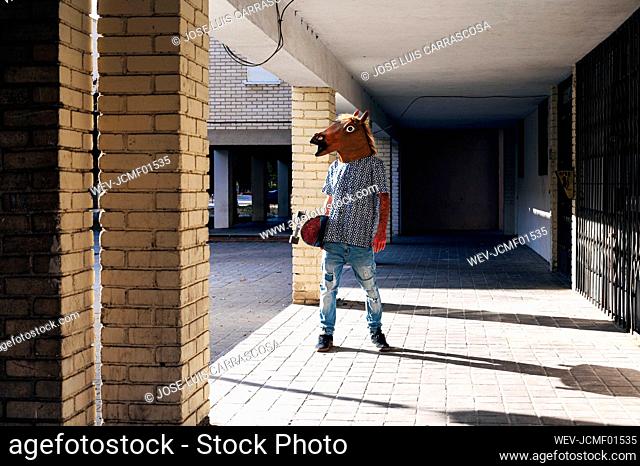 Man holding skateboard wearing horse mask while standing on footpath during sunny day
