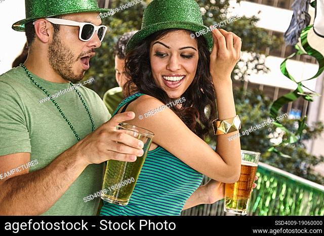 Young diverse couple dancing together wearing green sparkle hats and holding beer glasses for St. Patrick's Day