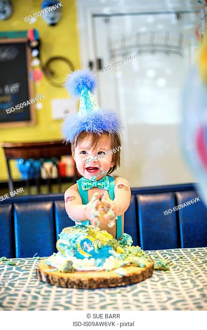Baby boy wearing party hat smashing first birthday cake at table
