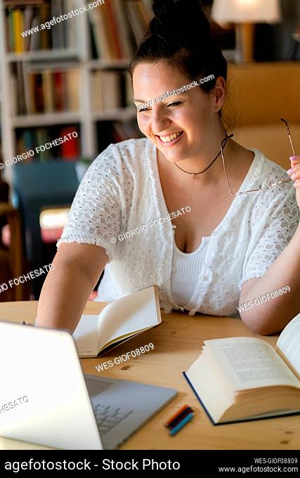 Smiling young woman studying over laptop on table in coffee shop